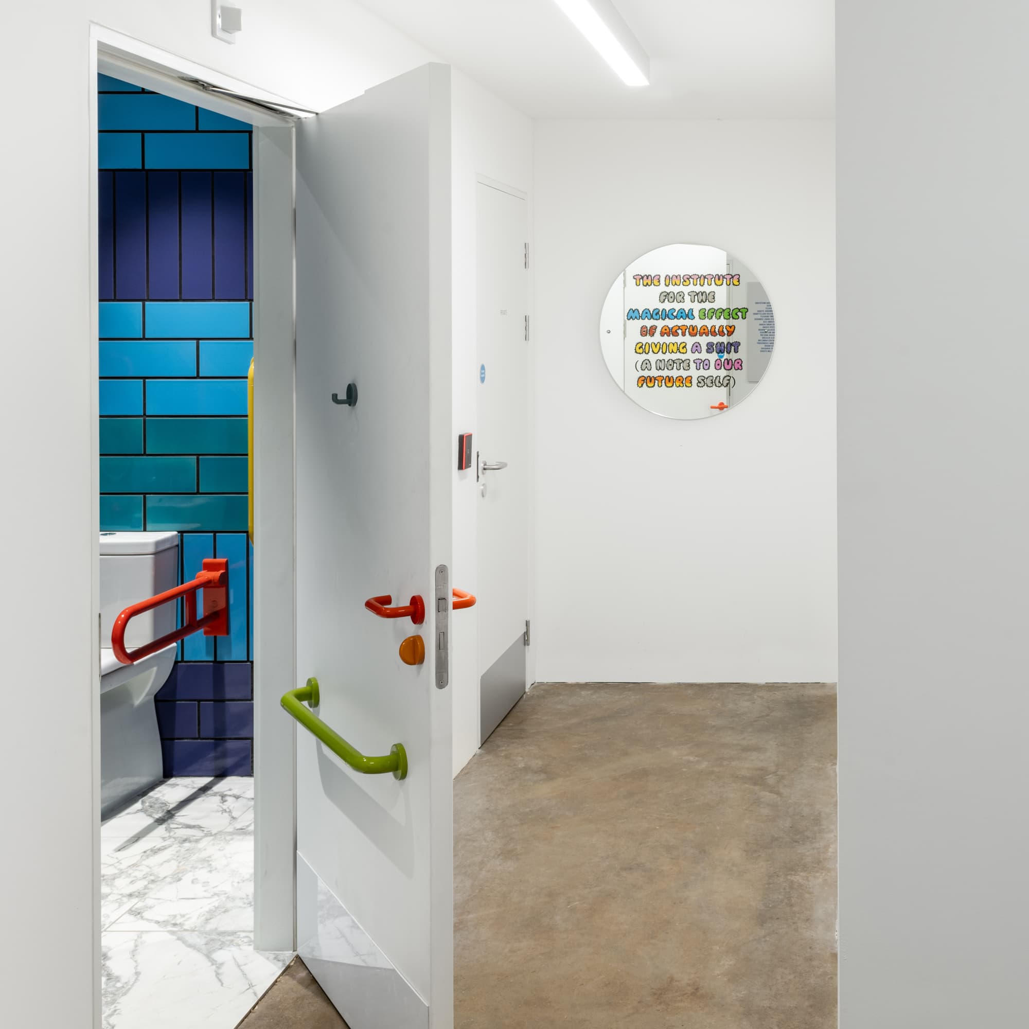 A photograph of a white corridor. A door is open to the toilet with blue and purple tiles. At the end of the corridor is a circular mirror with multi-coloured text saying 'The Institute for the magical effect of actually giving a shit (a note to our future self).'
    