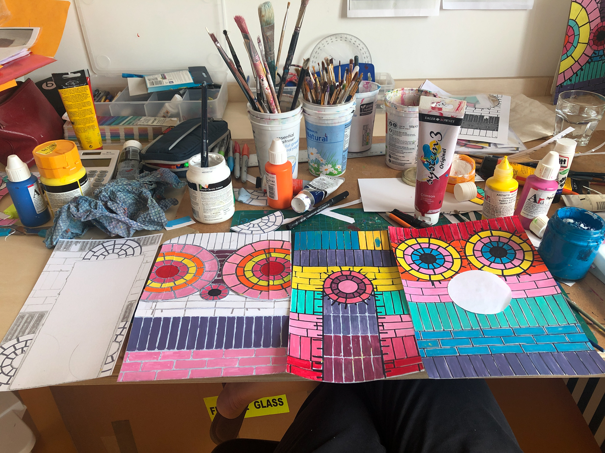 A photograph of a desk with 4 mock drawings of the tiled walls in pink, red, purple and blue. On the desk are several pots filled with paintbrushes, stacks of pens, and multiple tubes of paint.