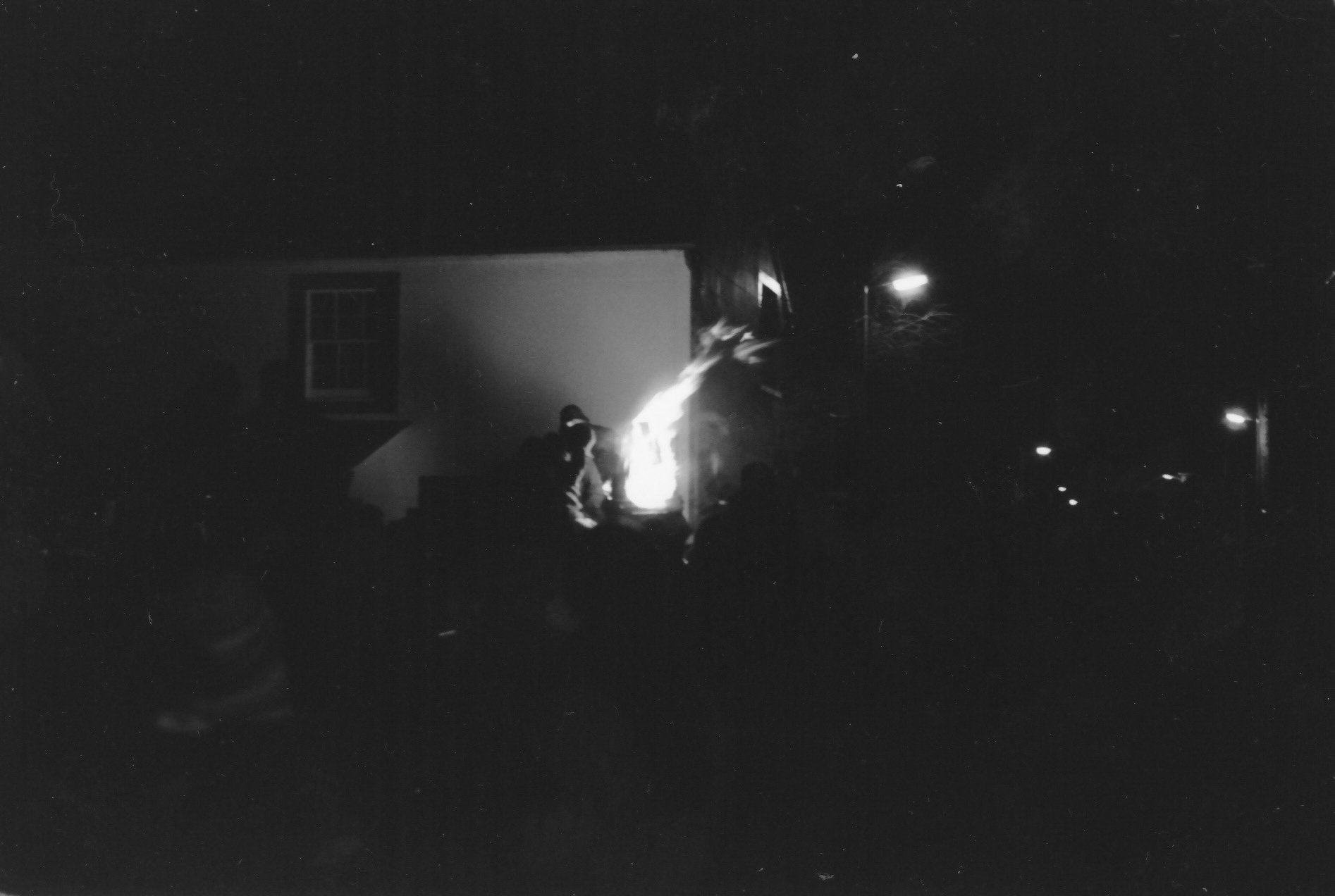 A blurry black and white photograph showing a figure standing next to a fire in front of a building, with street lights glowing in the dark.