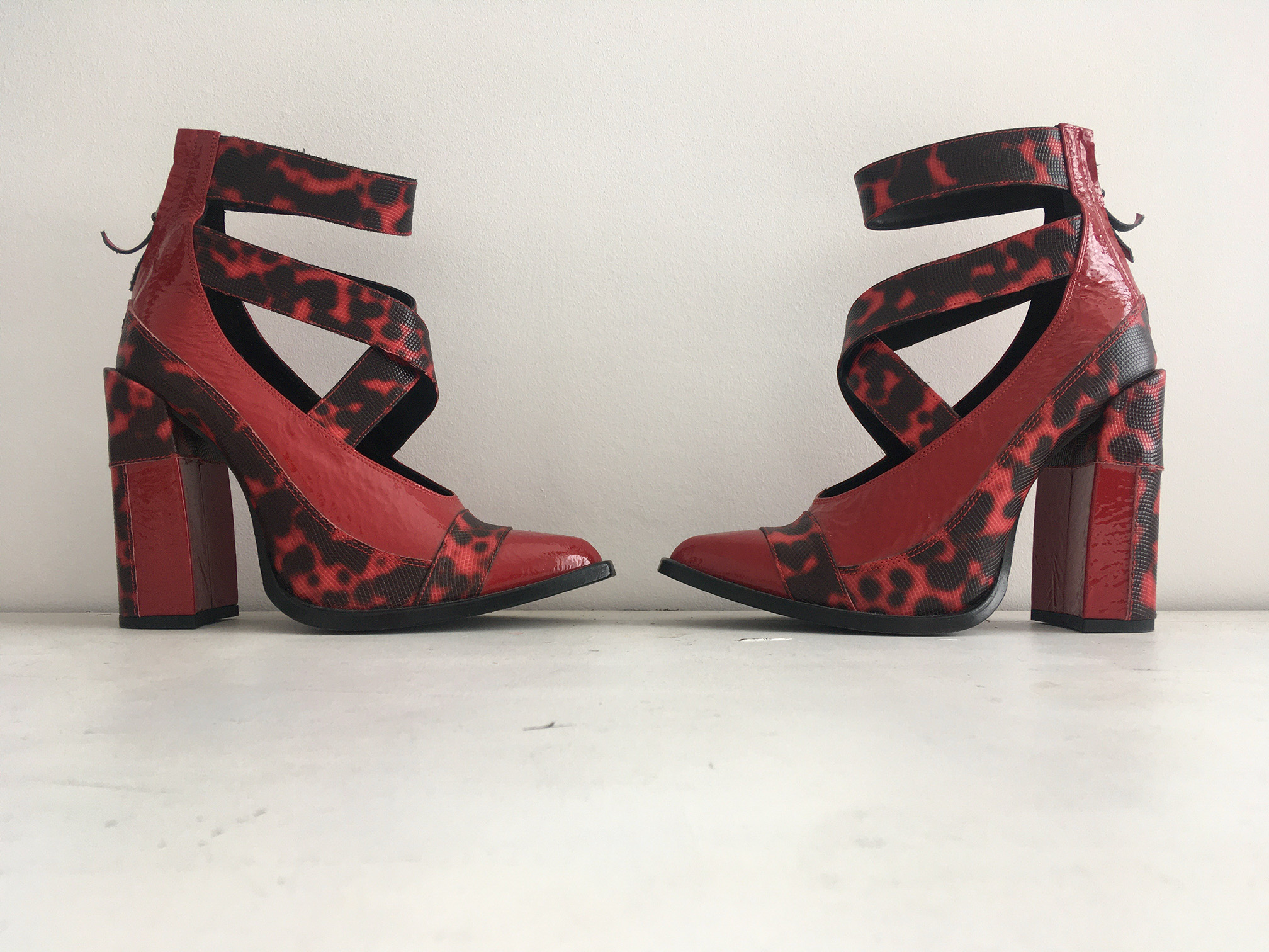 A photograph of 2 red high heeled shoes, facing each other against a white wall.