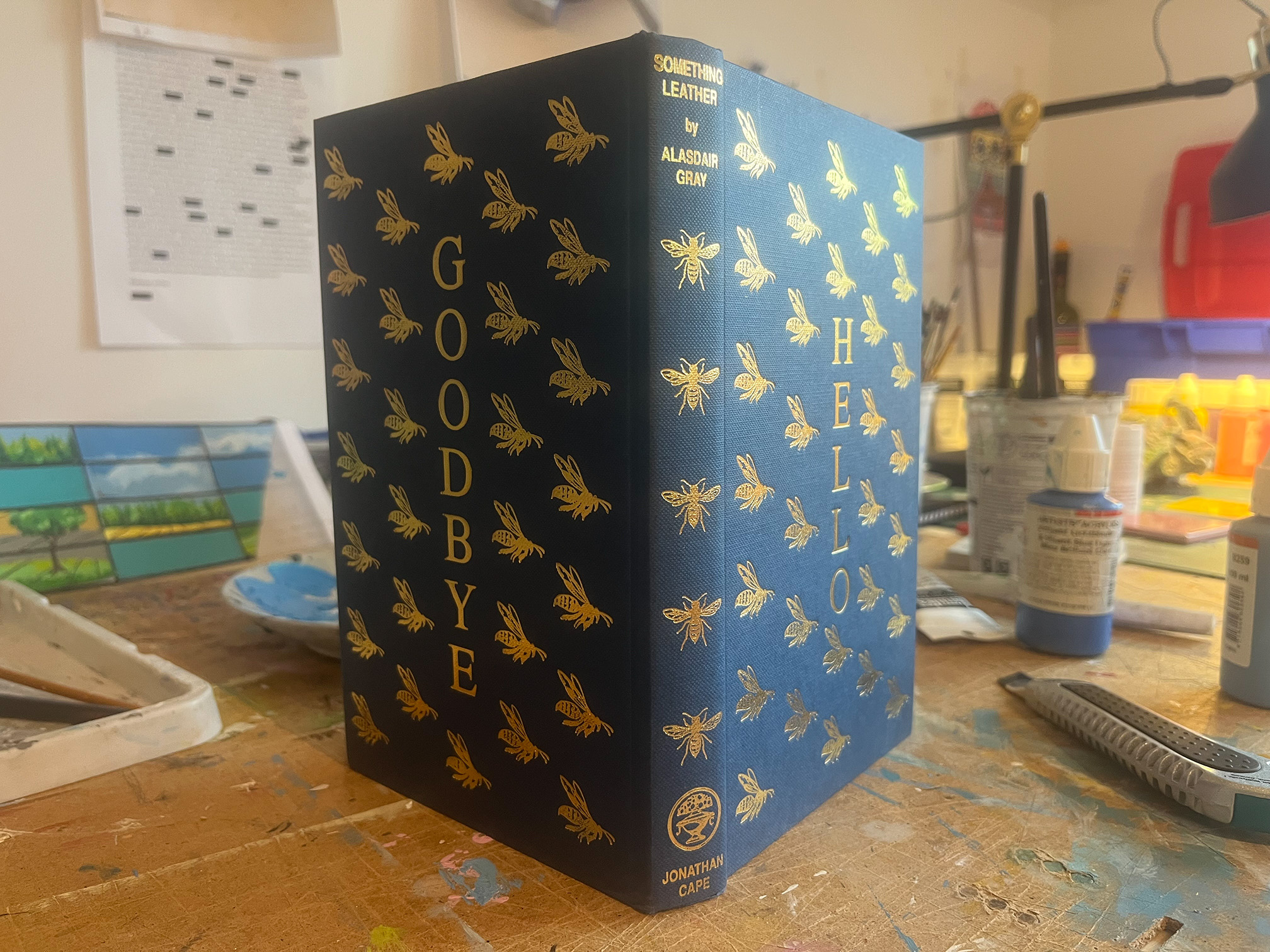 A photograph of the spine and cover of an open book standing upright on a brown desk. The blue book is covered in gold illustrations of bees, with the words GOODBYE and HELLO in gold type vertically on the back and front covers respectively. Behind the book there are paint tubes and paint brushes.