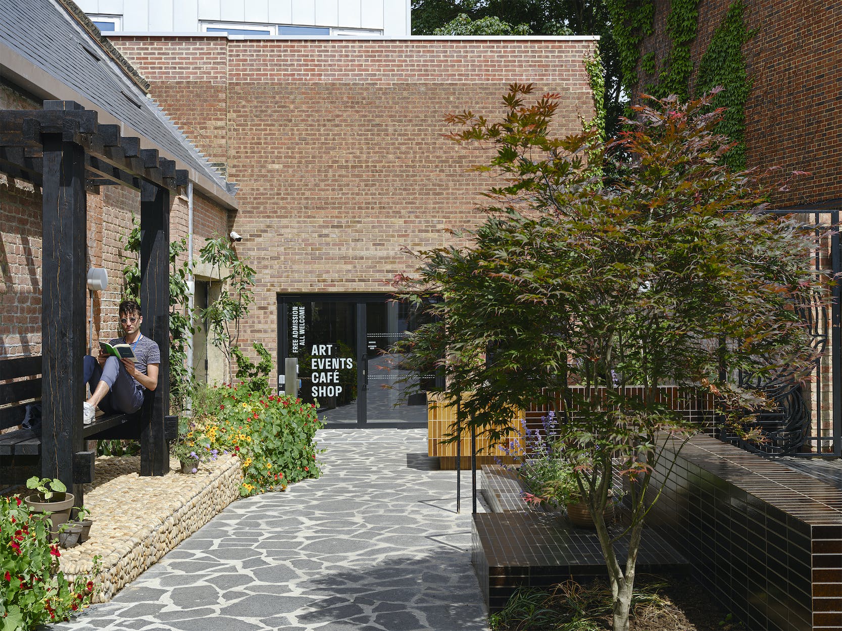 A photograph of the garden and entrance at Studio Voltaire. Grey slabs lead to the entrance doors of the bricked building. An acer tree stands on the right in front of brown tiles that make up steps and seating. On the left a man sits on a wooden pergola on a raised pebbled platform.