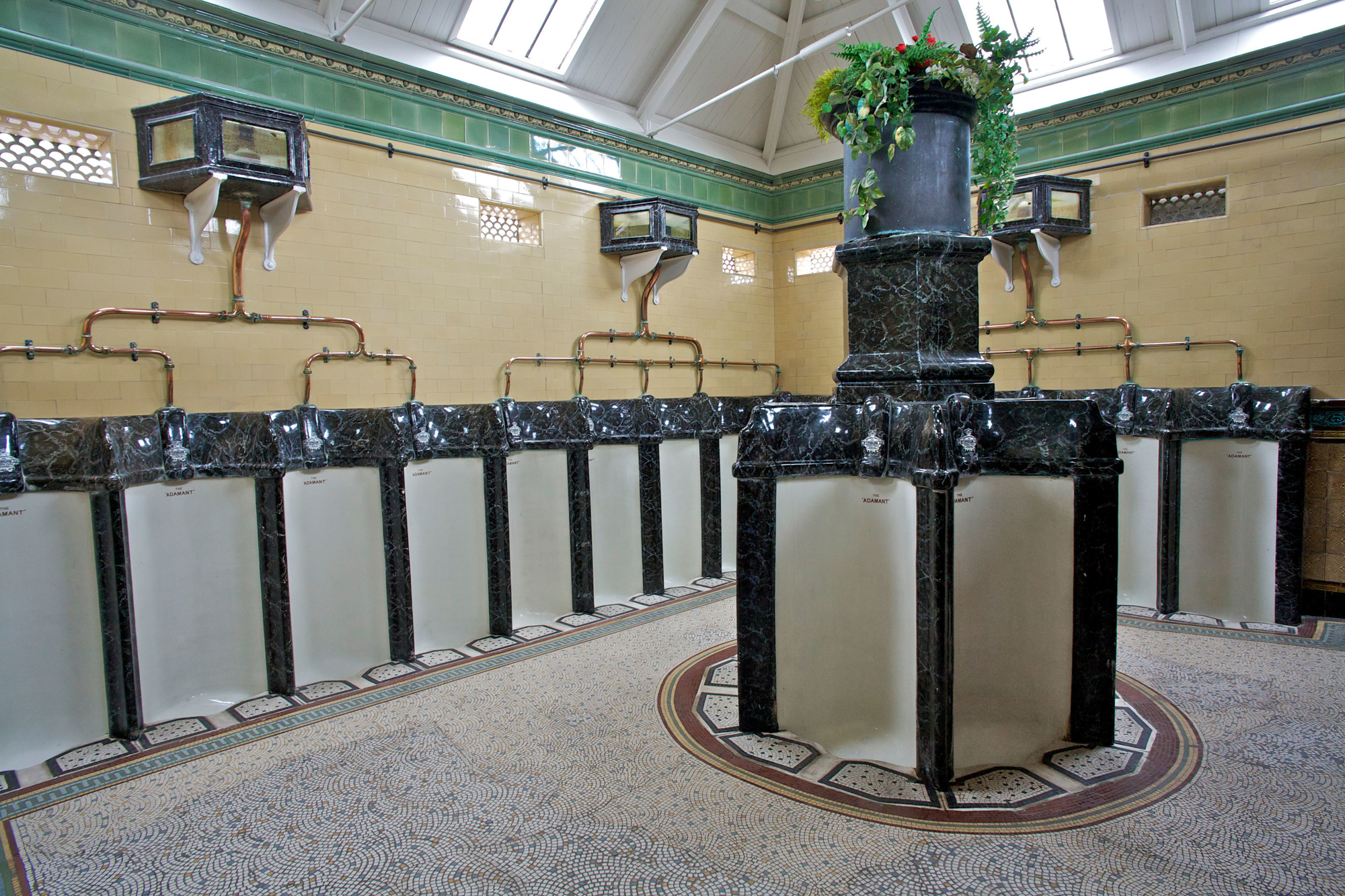 A photograph of a public bathroom with urinals along the walls and around a central pillar with a cascade of green plants on top. The urinals are white with black marble edges. Across the yellow tiled walls, are networks of copper pipes going upwards to black marble boxes. The floor in the photograph is covered in small white, red and blue tiles.   