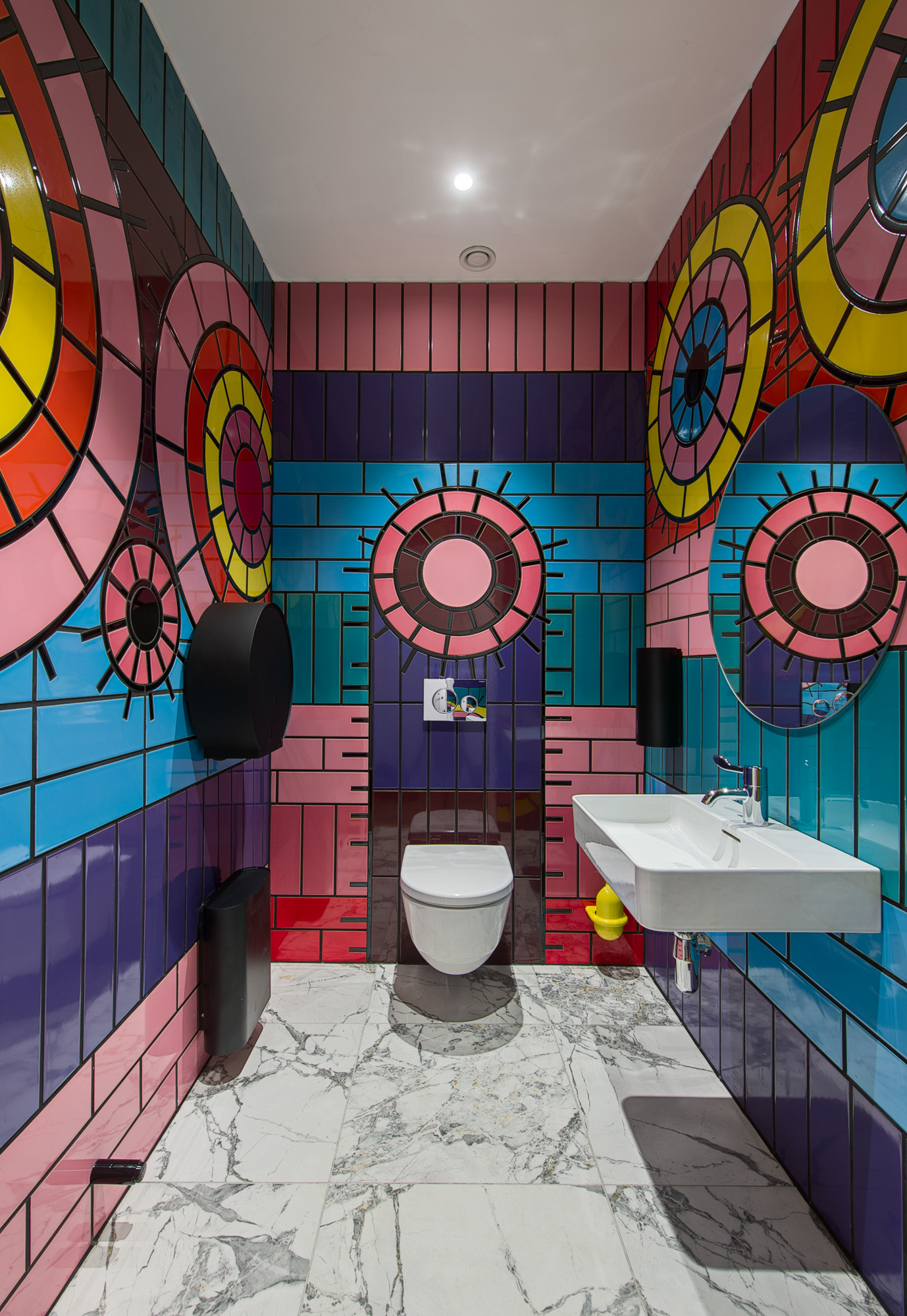 A photograph of a bathroom, with a view of 3 walls. The bathroom walls are covered in blue, purple and pink tiles in stripes, with pink and yellow circles of tiles. On the back wall is a white toilet with the seat down, with a square sink on the right wall, and a black toilet roll holder and sanitary bin on the left wall. The floor is white marble, and the ceiling is white.