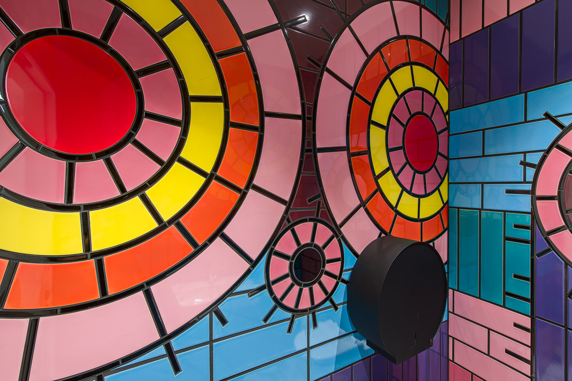 A photograph of 2 tiled walls with a black toilet roll holder on the left wall. The tiles form 2 large circles and one smaller circle, against horizontal stripes of coloured tiles. The larger circles are made of pink, yellow and orange tiles, with a red circle tile in the centre. The smaller circle is made of pink tiles with a black circle in the centre. All the circles have black tiles coming out of them in lines.