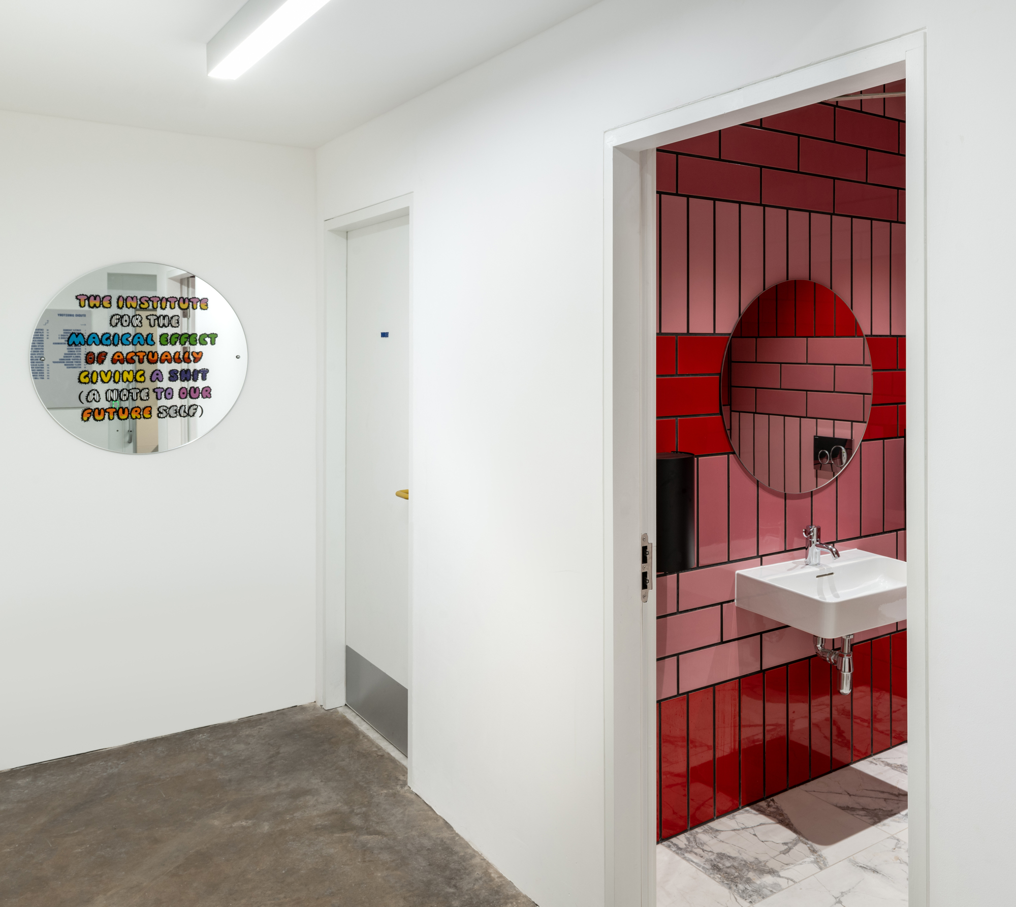 A photograph of a white corridor showing 2 white walls, a grey floor, and 2 doors. The first door is open to show a pink tiled wall with a square white sink and circular mirror. At the end of the corridor is a circular mirror with multi-coloured text saying 'The Institute for the magical effect of actually giving a shit (a note to our future self)'.